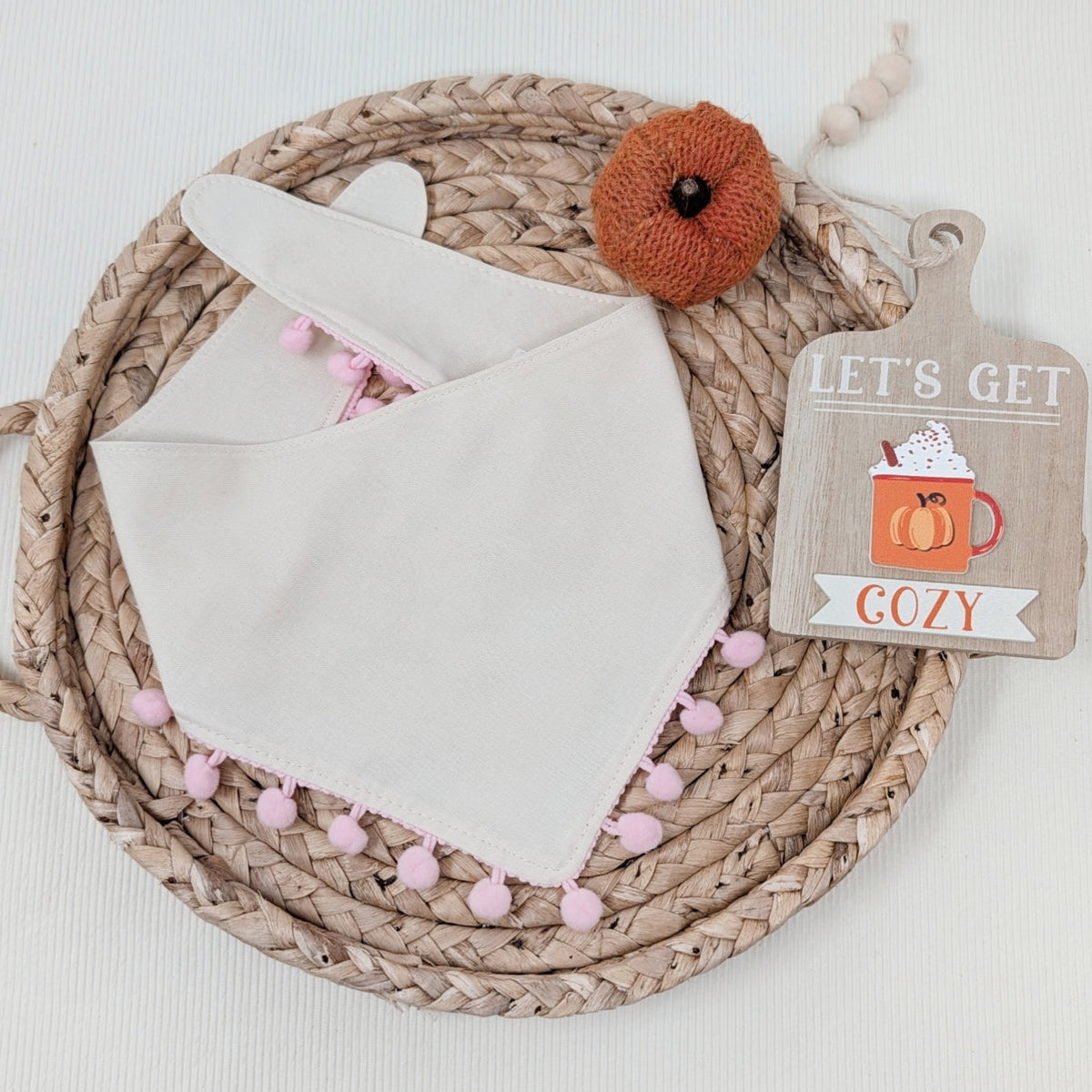 An off-white fabric bib with pink pom-pom trim, folded and displayed on a woven jute placemat. A small orange knit pumpkin decoration and a wooden tag that reads 'LET'S GET COZY' are placed beside it on a white background.