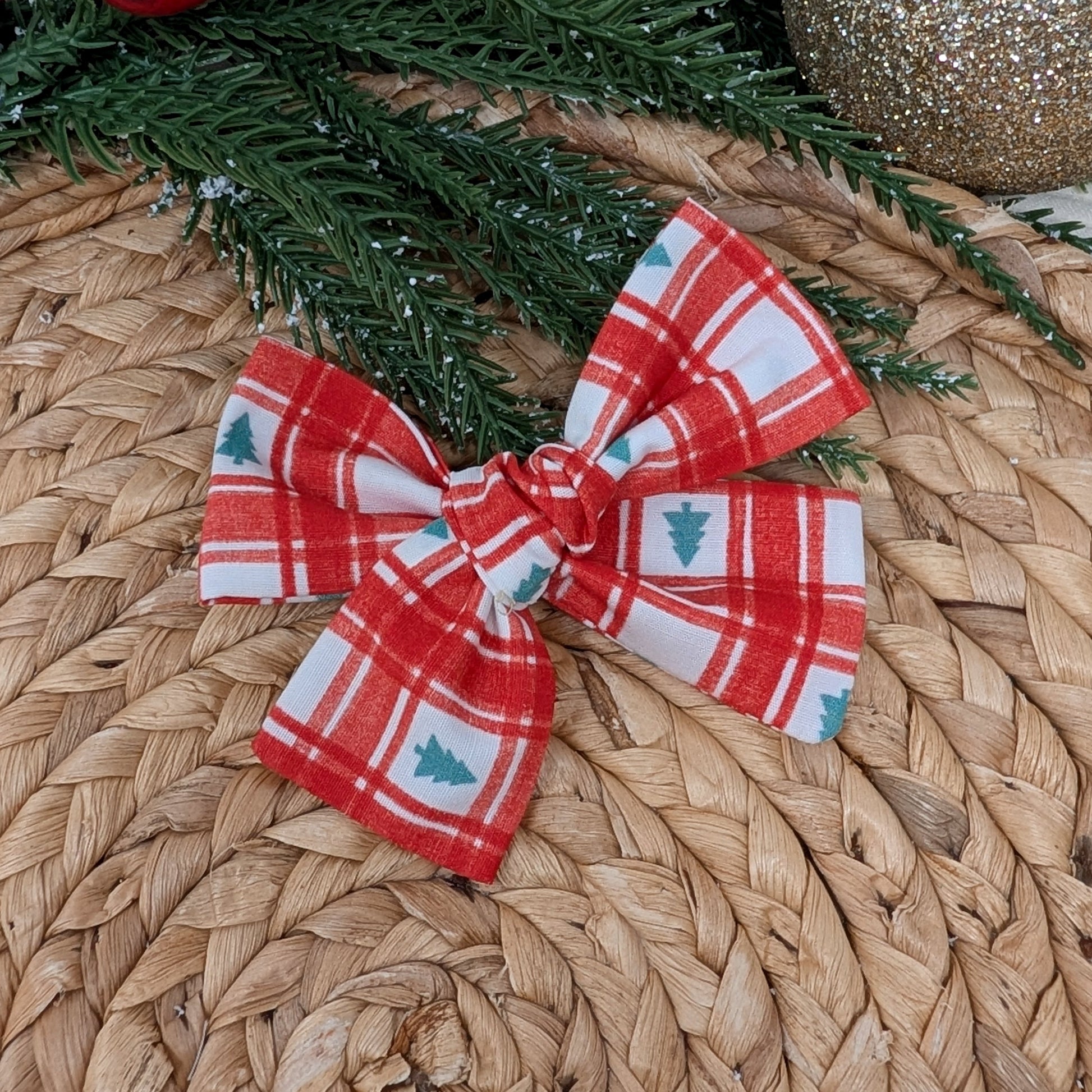 Red and white plaid bow with Christmas tree pattern on a woven surface with greenery and ornaments.
