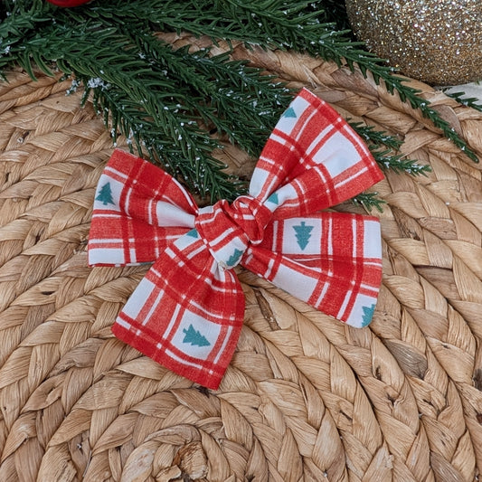 Red and white plaid bow with Christmas tree pattern on a woven surface with greenery and ornaments.
